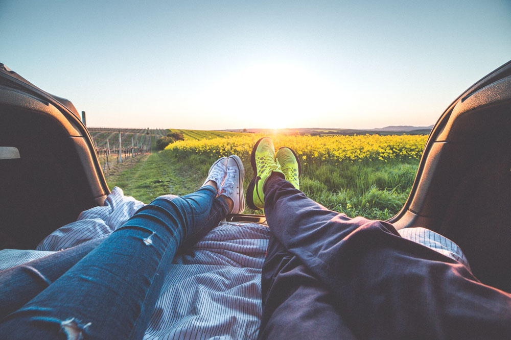 A romantic sunrise view with a man and woman resting on the green grass, their legs visible, as vibrant colors fill the sky.